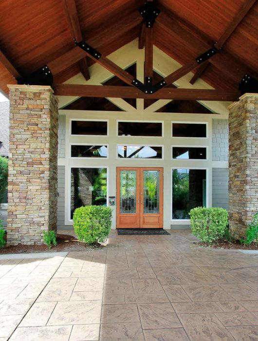 Entrance of a modern house with stone pillars, wooden double doors, and landscaped shrubs.